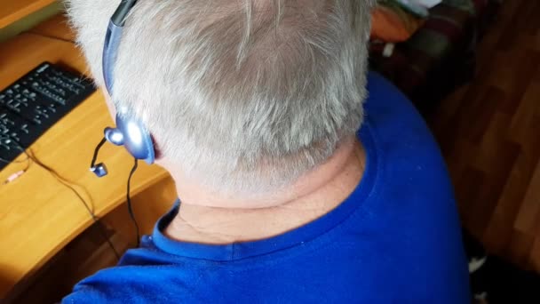 Siwy starzec ze słuchawkami korzysta z komputera do nauki na odległość, słucha muzyki i komunikuje się przez Skype 'a. Aktywny tryb życia emerytalnego, widok z tyłu — Wideo stockowe