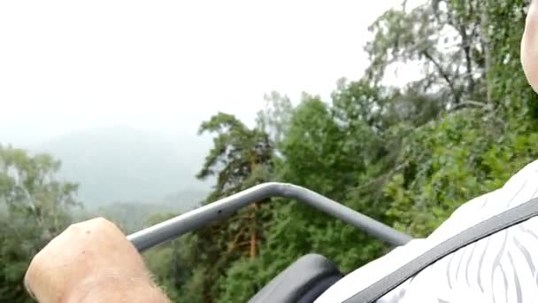 Een oudere, volwaardige mannelijke gepensioneerde zit in een kabelbaanstoel en onderzoekt het landschap met bos, bergen en mist, het concept van actieve recreatie — Stockvideo