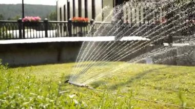 Yaz parkında çim sulama için modern fan sistemi, su makinesinin jetleri arasından bitkilerin görüntüsü, güneşte ışıldayan temiz su damlaları, HD video.