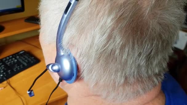 Un anziano dai capelli grigi con le cuffie utilizza un computer per l'apprendimento a distanza, ascolta musica e comunica via Skype. Stile di vita attivo pensionamento, vista dal retro — Video Stock
