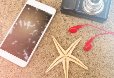 Plaj tatili ve seyahat konsepti, akıllı telefon, kamera, kulaklık ve kumsaldaki deniz yıldızı, akıllı telefon ekranına yansıyan güneş ışığı..