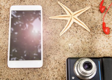 Plaj tatili ve seyahat konsepti, akıllı telefon, kamera, kulaklık ve kumsaldaki deniz yıldızı, akıllı telefon ekranına yansıyan güneş ışığı..