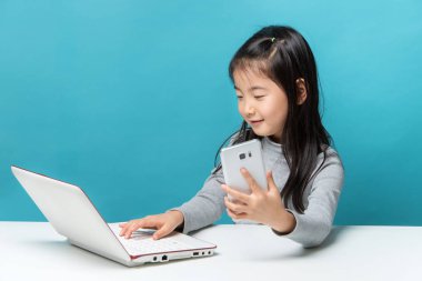 Sevimli Asya küçük kız onu beyaz laptop ve mavi içinde izole bir akıllı telefonlar ile masada oturuyor