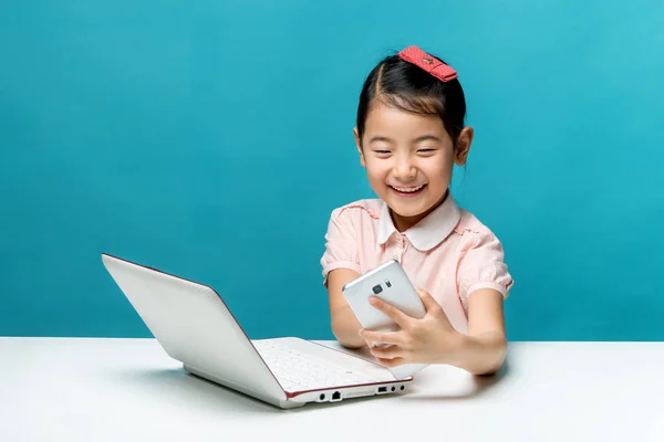 Χαριτωμένο Ασίας κοριτσάκι κάθεται στο τραπέζι με το laptop της λευκό και ένα smartphone, απομονωμένη πάνω μπλε — Φωτογραφία Αρχείου