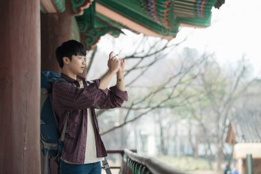 Genç bir adam bir Kore geleneksel evde bir sırt çantasıyla yolculuk yapıyor. Benim smartphone kullanarak fotoğraf çekiyorum.