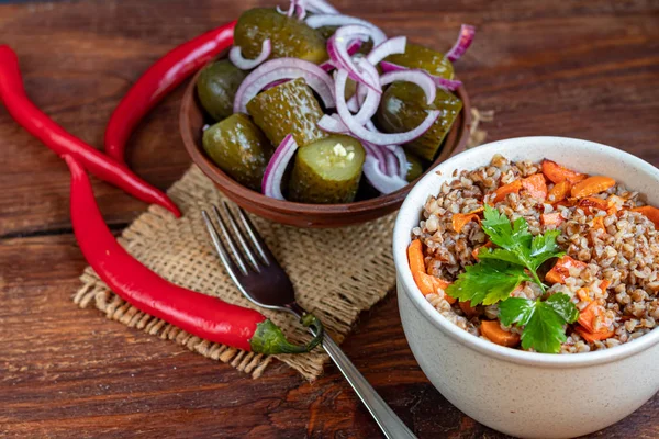 Boekweit pap in een kom met gestoofde wortelen. Op de achtergrond zijn gefermenteerde gepekelde komkommers en rode chili pepers. — Stockfoto