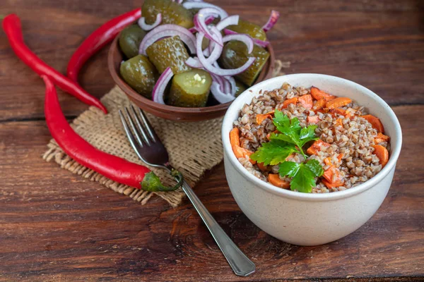 Boekweit pap in een kom met gestoofde wortelen. Op de achtergrond zijn gefermenteerde gepekelde komkommers en rode chili pepers. — Stockfoto