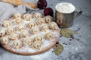 Ahşap bir tahtanın üzerinde pişmemiş, donmuş hamur tatlısı. Arka planda un, biber, yeşillik ve soğanın yanında hafif arkaplanda metal bir kupa var..