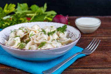 Yeşil soğanlı haşlanmış hamur tatlısı. Arka planda yeşiller, kırmızı biber ve defne yaprakları var. Şeffaf bir kaymak kabında.