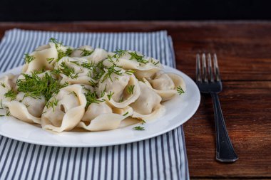 Yeşil soğanlı haşlanmış hamur tatlısı. Arka planda yeşiller, kırmızı biber ve defne yaprakları var. Şeffaf bir kaymak kabında.