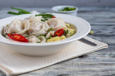 Soğan ve biberle süslenmiş köfteli çorba. Arka planda yeşiller, kırmızı biber ve defne yaprakları var. Hafif ahşap bir arka planda.