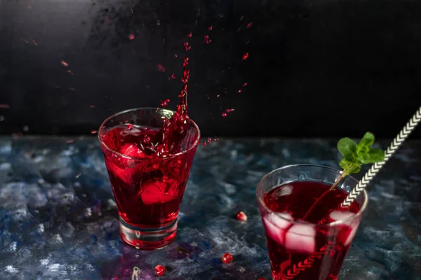 Jus de grenade dans des verres transparents. Vaporiser à partir d'un morceau de glace qui tombe. Sur un fond bleu sous une pierre. Tendance 2020. Les grains sont éparpillés à proximité . — Photo