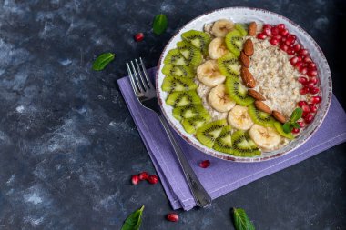 Yulaf ezmesi, fındık ve meyvelerden oluşan kahvaltı. Kivi portakallı nar badem muzlar bir tabak süsler. Sağlıklı beslenme, karanlık bir arka planda..