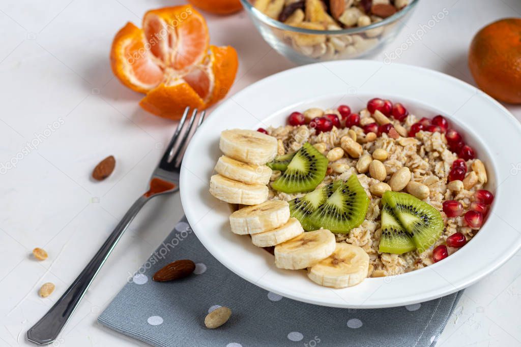 Breakfast consisting of oatmeal, nuts and fruits. Kiwi, banana, pomegranate and almonds decorate the plate. Healthy food, on a white background.