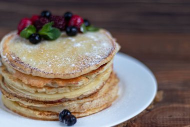 Ahududu, böğürtlen ve yabanmersinli bir yığın lezzetli krep. Ahşap bir arka planda. Pudra şekeri serpilmiş ve nane yapraklarıyla süslenmiş..