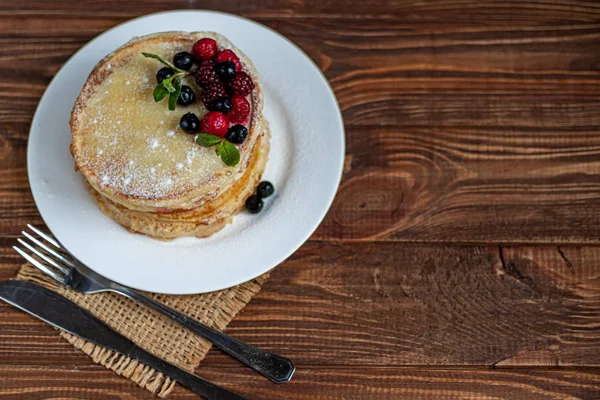 Uma pilha de deliciosas panquecas com framboesas, amoras e mirtilos. Em um fundo de madeira. Polvilhado com açúcar em pó e decorado com folhas de hortelã . — Fotografia de Stock