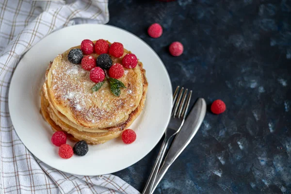 Una pila de deliciosos panqueques con frambuesas, moras y arándanos. Sobre un fondo de piedra oscura. Espolvoreado con azúcar glaseado y decorado — Foto de Stock