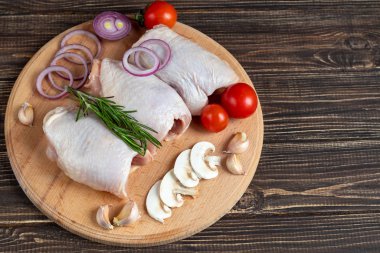 On a light wooden board, fresh skinless chicken thighs, next to mushrooms, with tomatoes, red onions and rosemary. Recipe of dish. Ingredients.