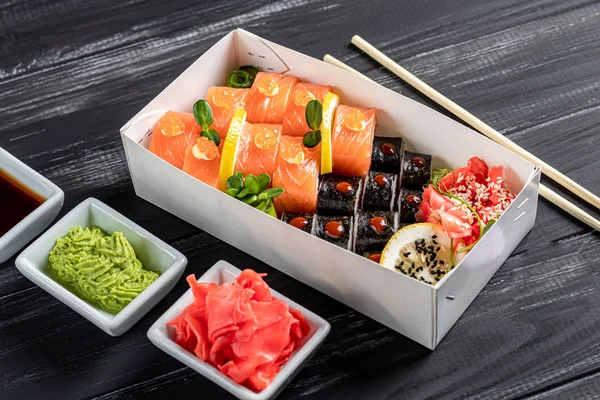 Krabici sushi Nigiri, Uramaki California, Philadelphia, na černém kamenném talíři. Sushi menu v bílém transportním boxu na dřevěném pozadí. — Stock fotografie