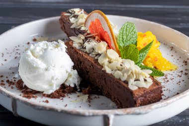 Brownie çikolatalı tatlı, bir kepçe beyaz dondurma, bir dilim mango ve bir dilim greyfurt. Arka planda bir kapuçino var. Karanlık bir arka planda.
