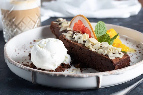 Brownie-Schokolade-Dessert mit einer Kugel weißem Eis, Mango-Scheiben und einer Scheibe Grapefruit. Im Hintergrund steht ein Cappuccino. auf dunklem Hintergrund. — Stockfoto