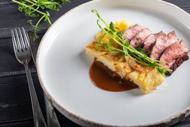 Yanında patates güveci olan ördek göğsü. Bezelye mikroyeşili, domates ve biber soslu. Beyaz bir tabakta. Ahşap bir arka planda,