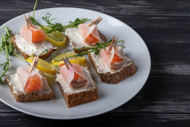 Sandwiches with salmon on dark bread, spread with cheese sauce. The meat is wrapped in a slice of daikon and fixed with a decorative clothespin. On a wooden background.