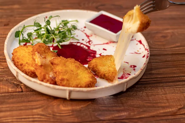 Camembert cheese fried in batter and breadcrumbs with cranberry sauce. Soft and viscous inside. The plate is decorated with a spray of sauce and microgreen. On a wooden background.