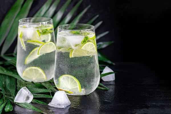 Mojito in a large transparent glass with lime and mint leaves and ice cubes. In the background are palm branches. On a black wooden background. — Φωτογραφία Αρχείου