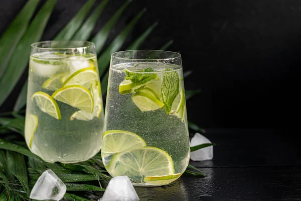 Mojito in a large transparent glass with lime and mint leaves and ice cubes. In the background are palm branches. On a black wooden background. — Stok fotoğraf