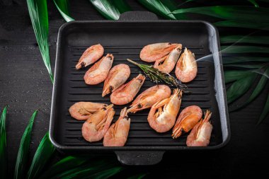 Grilled prawns in a pan with a sprig of rosemary for frying. On the substrate are green palm leaves. On a black wooden background.