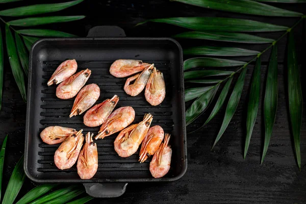 Grilled prawns in a pan with a sprig of rosemary for frying. On the substrate are green palm leaves. On a black wooden background. — Stock Photo, Image