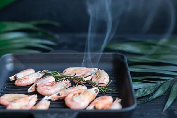 Grilled prawns in a pan with a sprig of rosemary for frying. On the substrate are green palm leaves. On a black wooden background. — Stock Photo, Image