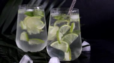 Büyük şeffaf bir bardakta mojito, kireç, nane yaprağı ve buz küpleriyle. Arka planda palmiye dalları var. Siyah tahta bir arka planda. Boşluğu kopyala