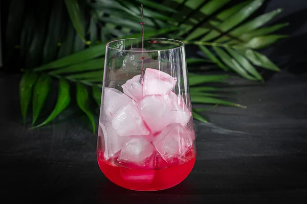 Raspberry-barberry drink in a transparent glass with ice. The drink is poured into a glass. Added palm branches and raspberries. Black background. — Stock fotografie
