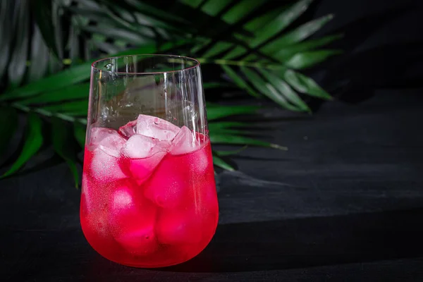 Малина-барбарис напиток в прозрачном стакане со льдом. Напиток наливают в стакан. Добавлены пальмовые ветви и малина. Чёрный фон . — стоковое фото