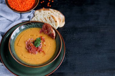 Ahşap arka planda mercimek çorbası. Dilimlenmiş pastırma ve bitkilerle servis edilir. Ciabatta parçaları yakınlarda. Arka planda ham büyümeler var. Boşluğu kopyala