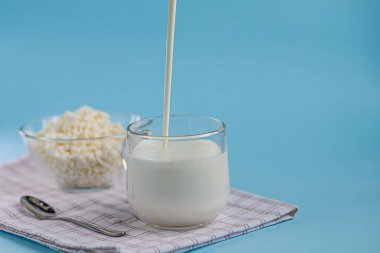 Şeffaf bir kasede şeffaf bir kaşık ve bir bardak kefir akıntısının mavi arka plana döküldüğü taze süzme peynir. Mayalanmış yiyecekler. Sağlık için iyi bir bakteri. Boşluğu kopyala