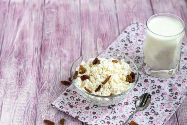 Şeffaf bir kasede kuru üzümlü taze süzme peynir. Arka planda bir bardak kefir var. Mayalanmış yiyecekler. Sağlık için iyi bir bakteri. Hafif arkaplan. Boşluğu kopyala