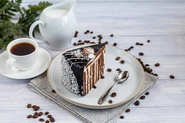 A slice of zebra cake with chocolate icing. On a light background. In the background is a cup of coffee. Coffee beans are scattered throughout. Copy space
