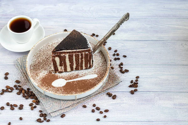 A slice of zebra cake with chocolate icing. On a light background. In the background is a cup of coffee. Coffee beans are scattered throughout. Copy space