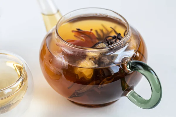 Blooming black tea in a glass teapot. On a light surface. Nearby are tea briquettes of leaves and flowers. In the background is a cake. Copy space