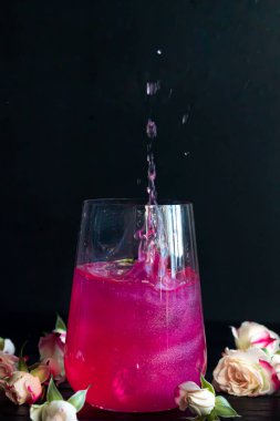 Bardaktaki parlak ahududu içeceği buzlu ahşap bir masanın üzerinde duruyor. Arka planda güller ve bir çelenk var. Boşluğu kopyala