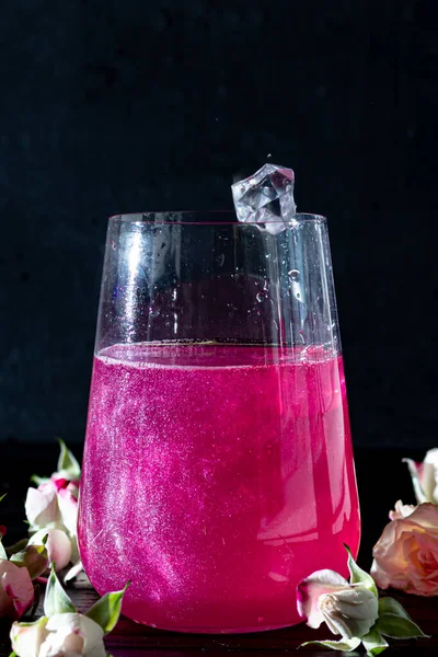 一个闪闪发光的覆盆子饮料在一个玻璃杯站在一个木制桌子上 上面有冰块 背景是玫瑰和花环 复制空间 — 图库照片