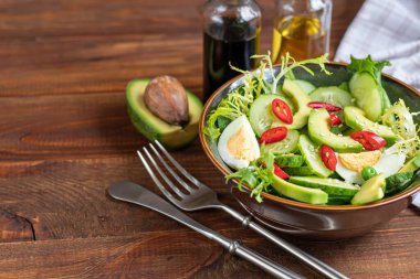 Taze salatalık, ıspanak yaprağı, roka, avokado. Dilimlenmiş yumurta ve kırmızı biberle servis ediliyor. Beslenme düzeni. Tüm aile için bir kahvaltı. Vitamin tabağı. Koyu ahşap bir arka planda. Boşluğu kopyala