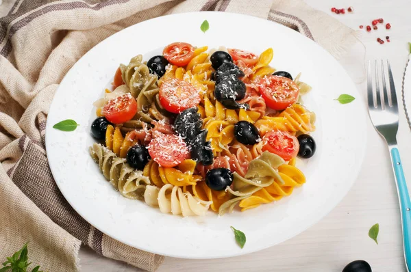 Massa colorida com azeitonas, tomates (cereja), manjericão e queijo em um prato branco em um fundo de madeira branco. Perto de talheres. Cozinha Italiana — Fotografia de Stock