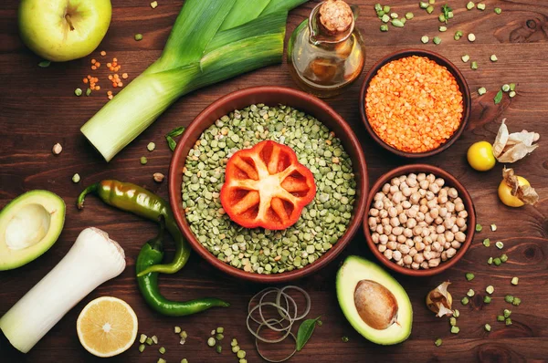 Diverse, multi-colored bean grains, such as peas, chickpeas and lentils in a clay plate on a brown wooden background. Nearby are vegetables such as onion, hot peppers, onions, avocado and physalis. Vegetarian, vegan concept — Stock Photo, Image