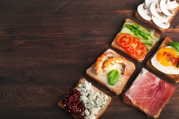 Sandwichs juteux lumineux avec différentes garnitures telles que crevettes, fromage bleu, tomates séchées au soleil, concombres, œufs frits, jambon et champignons sur un fond en bois brun. Espace pour le texte. Manger fait maison — Photo