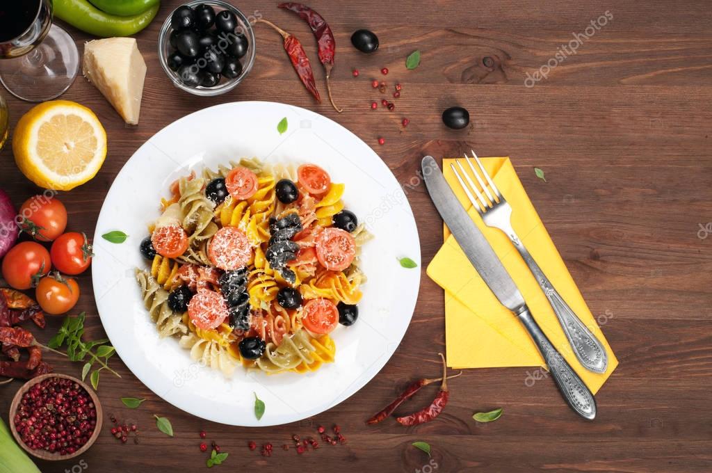 Classic Italian multicolored pasta (Fusilli) with cherry tomatoes, olives and cheese on a white plate on a brown wooden background. Near spices and condiments, glass of wine, vegetables. The recipes of the Italian cuisine (the concept)