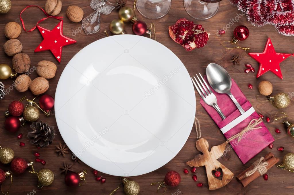 Empty white plate (to accommodate any festive dishes) near the Christmas toys, walnuts, anise stars and cinnamon, and other attributes of the New Year and Christmas on brown wooden background. The concept of supply of festive dishes (Serving holiday 